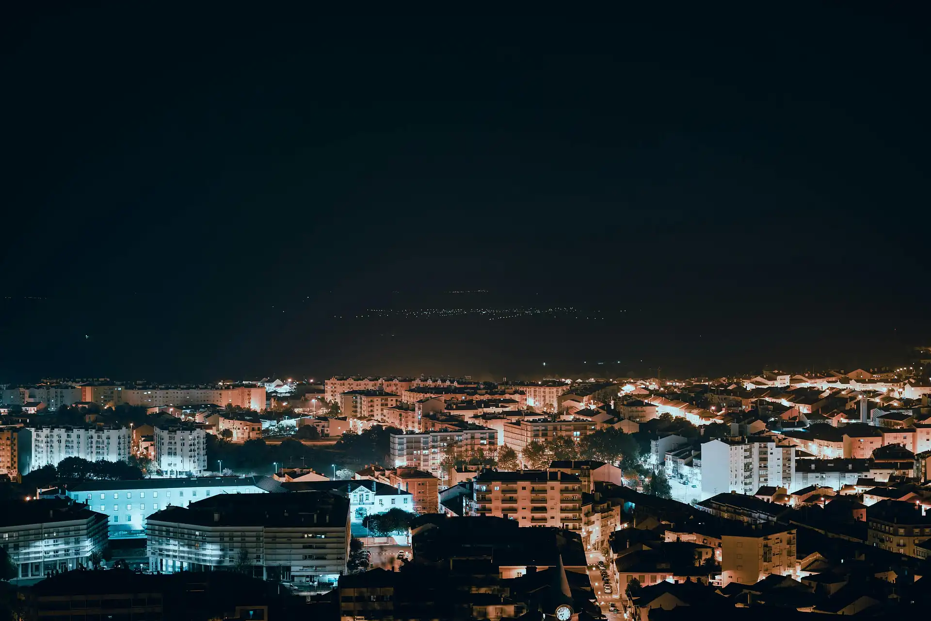 An aerial view of a city at night with teal and amber lights stretching to the horizon