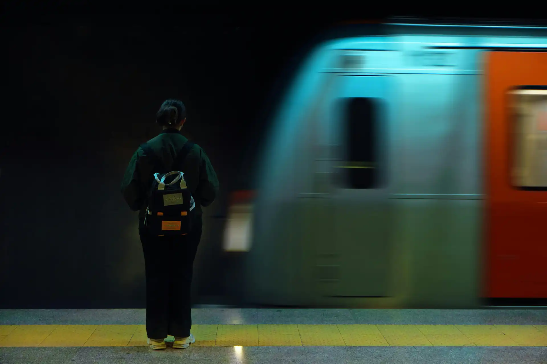 A person standing still on a subway platform as a motion blurred teal and orange train passes by