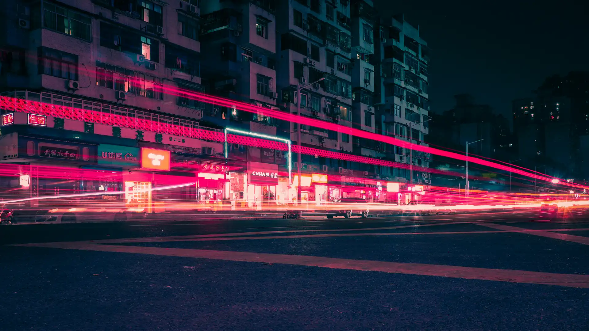 Red and pink light trails streaking across a dark urban street at night with illuminated building facades