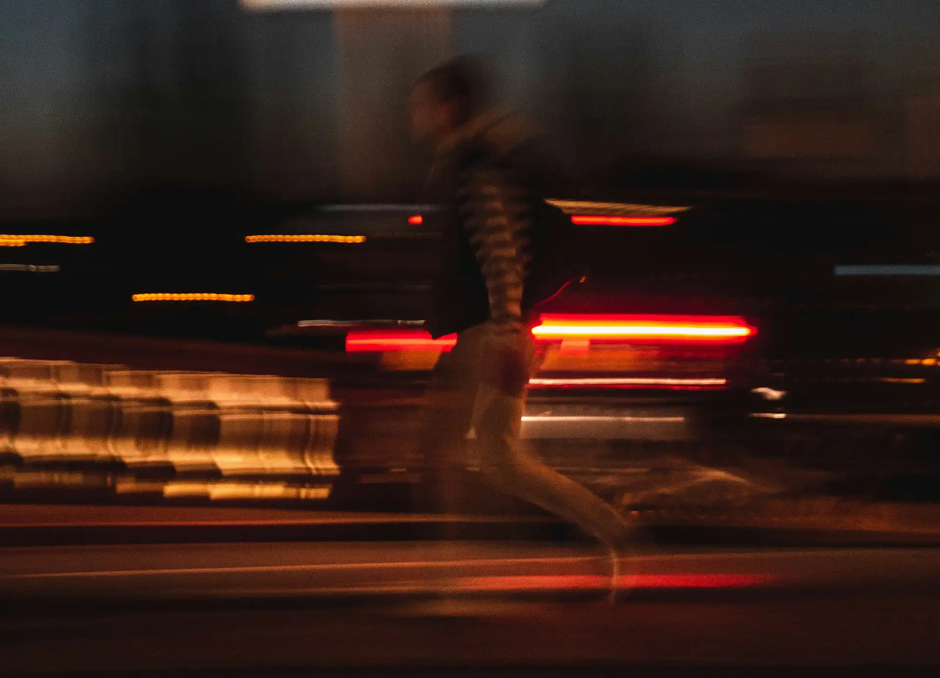 A blurred figure running through a dark street with red and amber car lights streaking in the background